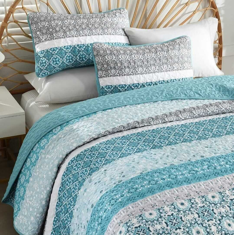 Bed with blue and gray patterned quilt and pillows in a room with a lamp and plant.