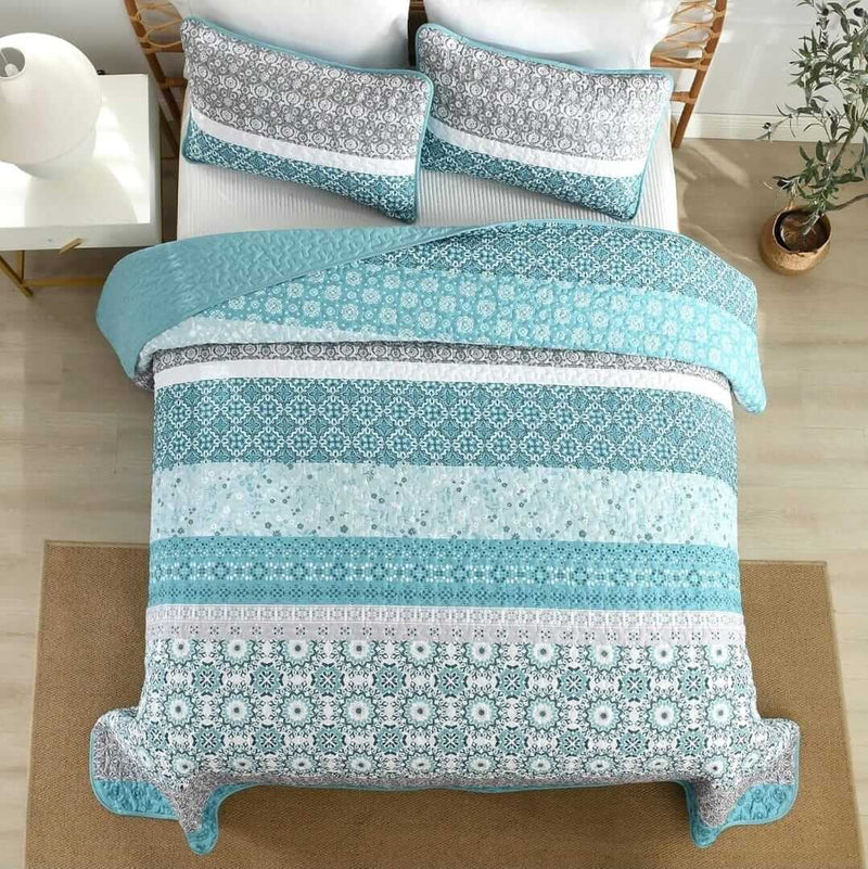 Bed with blue and gray patterned quilt and pillows in a room with a lamp and plant.