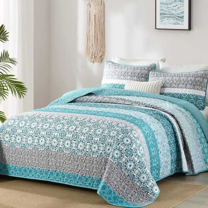 Bed with blue and gray patterned quilt and pillows in a room with a lamp and plant.