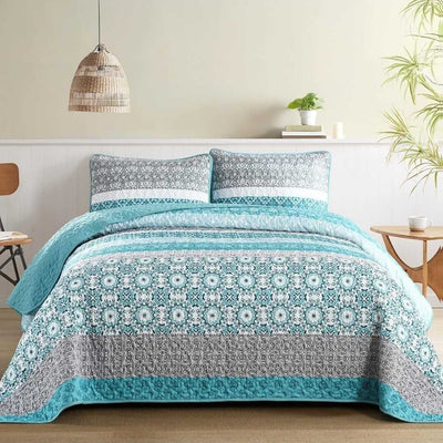 Bed with blue and gray patterned quilt and pillows in a room with a lamp and plant.