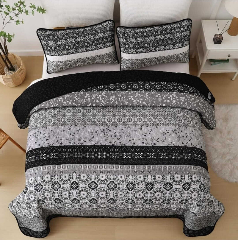 A colorful floral patterned cotton bedspread set on a bed, with a variety of matching and contrasting pillows, in a room with wooden furniture and a lamp.
