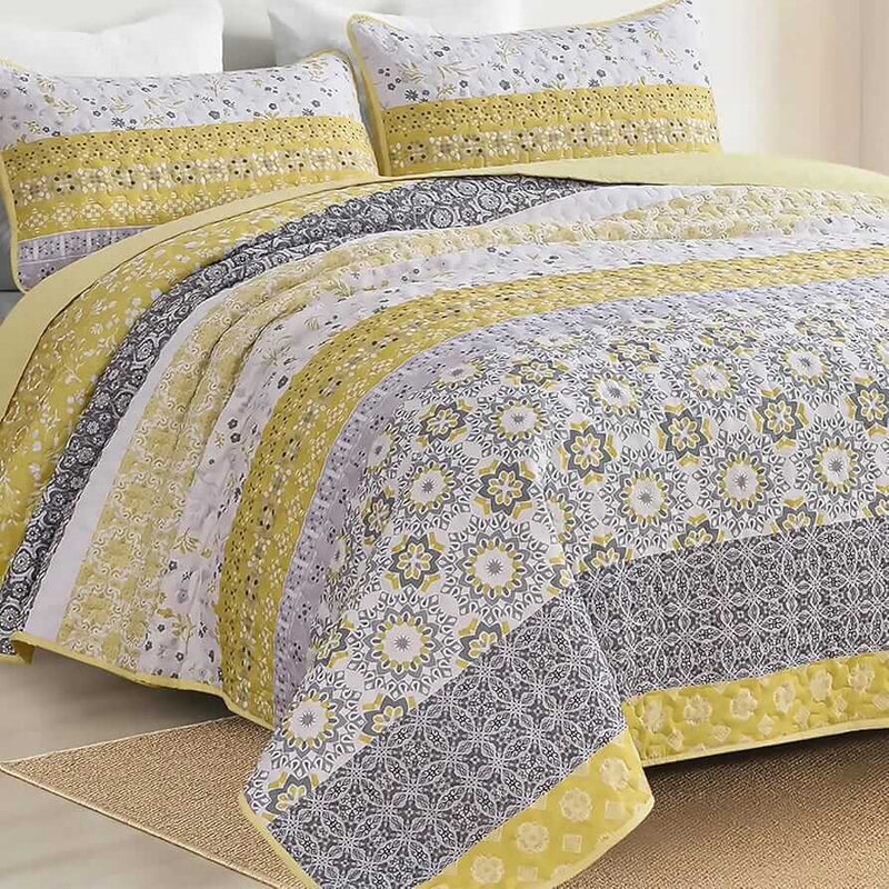 A colorful floral patterned cotton bedspread set on a bed, with a variety of matching and contrasting pillows, in a room with wooden furniture and a lamp.