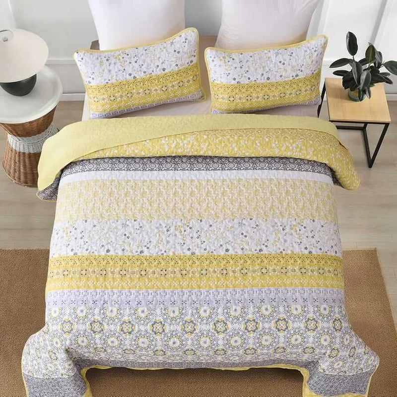 A colorful floral patterned cotton bedspread set on a bed, with a variety of matching and contrasting pillows, in a room with wooden furniture and a lamp.