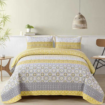 A colorful floral patterned cotton bedspread set on a bed, with a variety of matching and contrasting pillows, in a room with wooden furniture and a lamp.