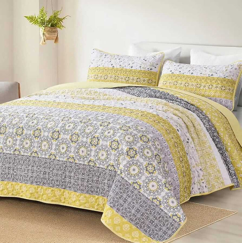 A colorful floral patterned cotton bedspread set on a bed, with a variety of matching and contrasting pillows, in a room with wooden furniture and a lamp.