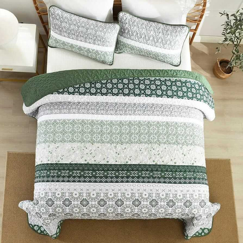 A colorful floral patterned cotton bedspread set on a bed, with a variety of matching and contrasting pillows, in a room with wooden furniture and a lamp.