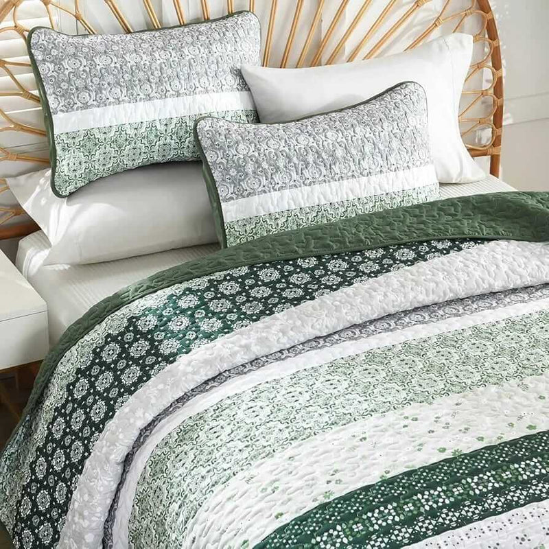 A colorful floral patterned cotton bedspread set on a bed, with a variety of matching and contrasting pillows, in a room with wooden furniture and a lamp.