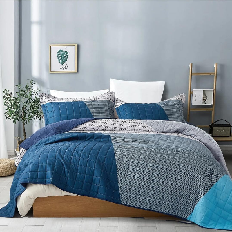 Blue and gray quilted bedding set on a bed in a room with a plant and wall art.