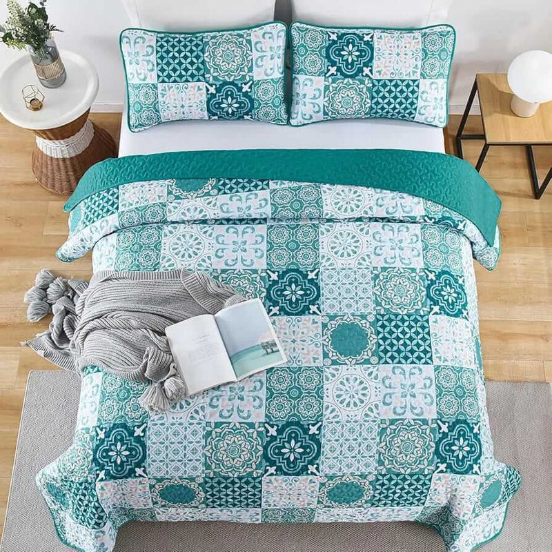 A colorful floral patterned cotton bedspread set on a bed, with a variety of matching and contrasting pillows, in a room with wooden furniture and a lamp.