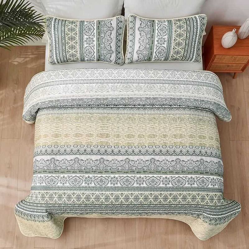 Patterned quilt set on a bed in a bedroom with a nightstand and plant.