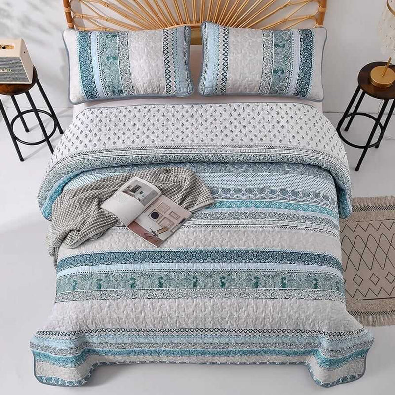 Patterned quilt set on a bed in a bedroom with a nightstand and plant.