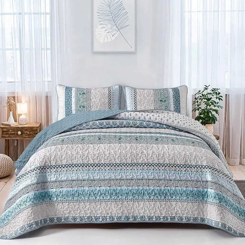 Patterned quilt set on a bed in a bedroom with a nightstand and plant.